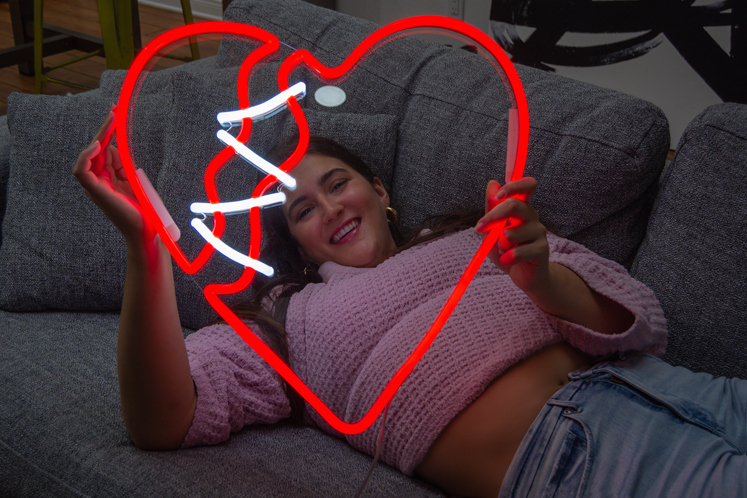Girl holding a red heart shaped neon sign while laying on a sofa