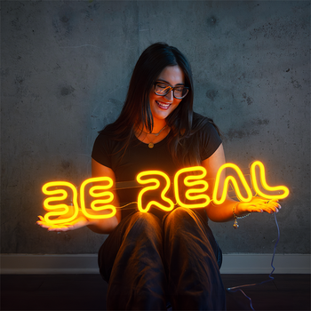girl holding a yellow neon sign that says "be real"