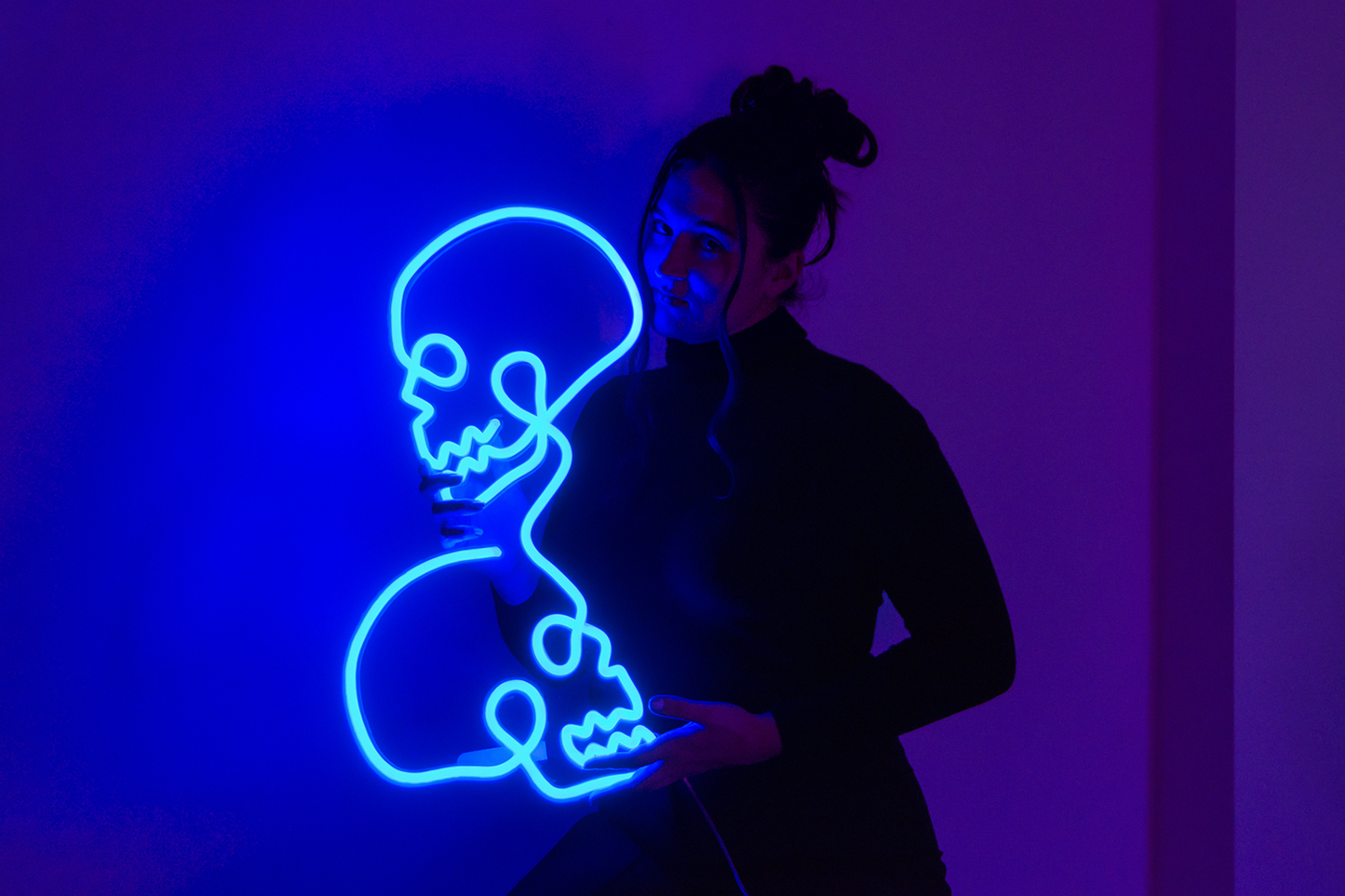 A pretty girl holding a blue neon sign of two skulls in a room with a blue and purple background