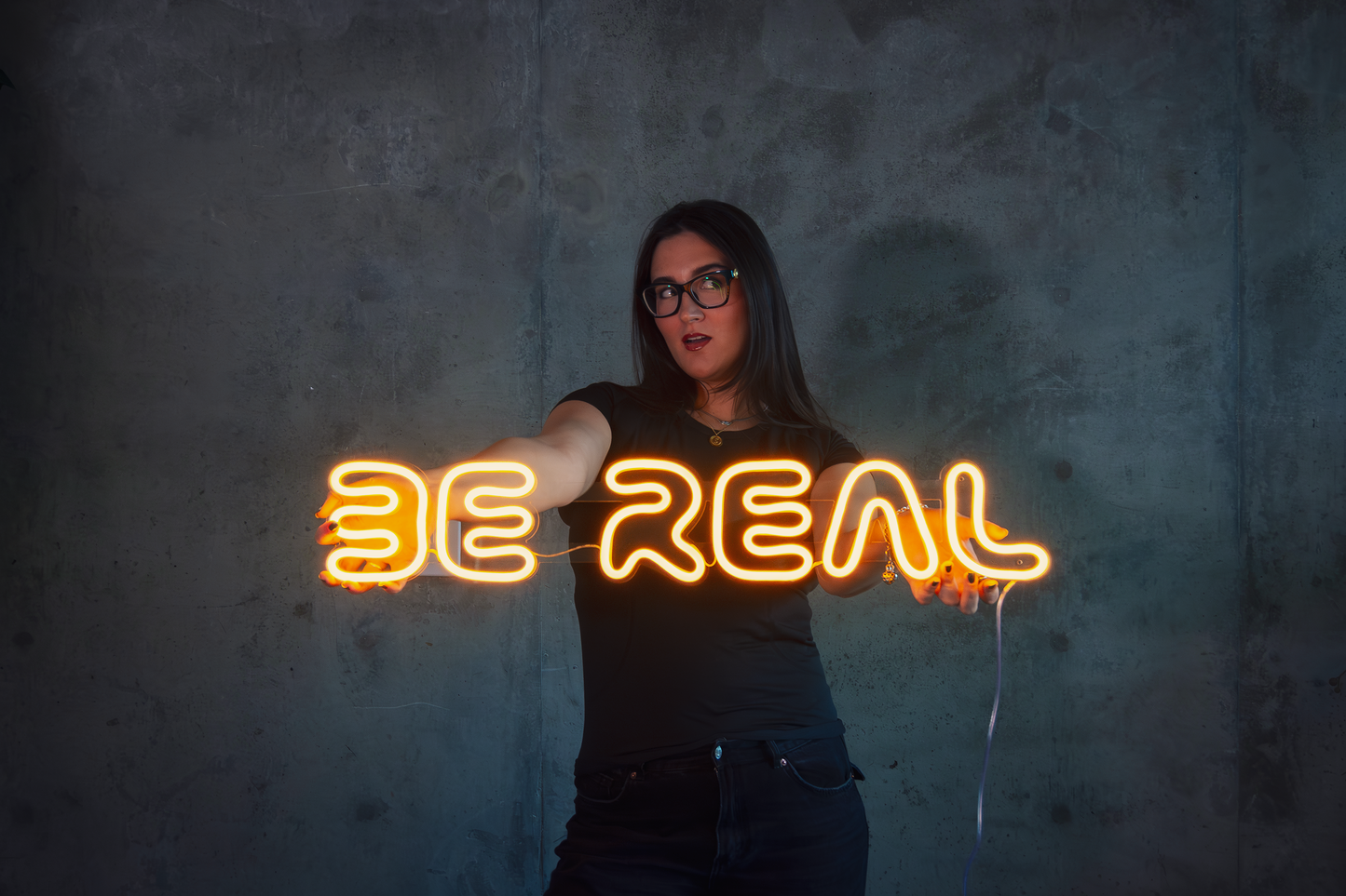 Girl holding a yellow neon sign that says "be real"