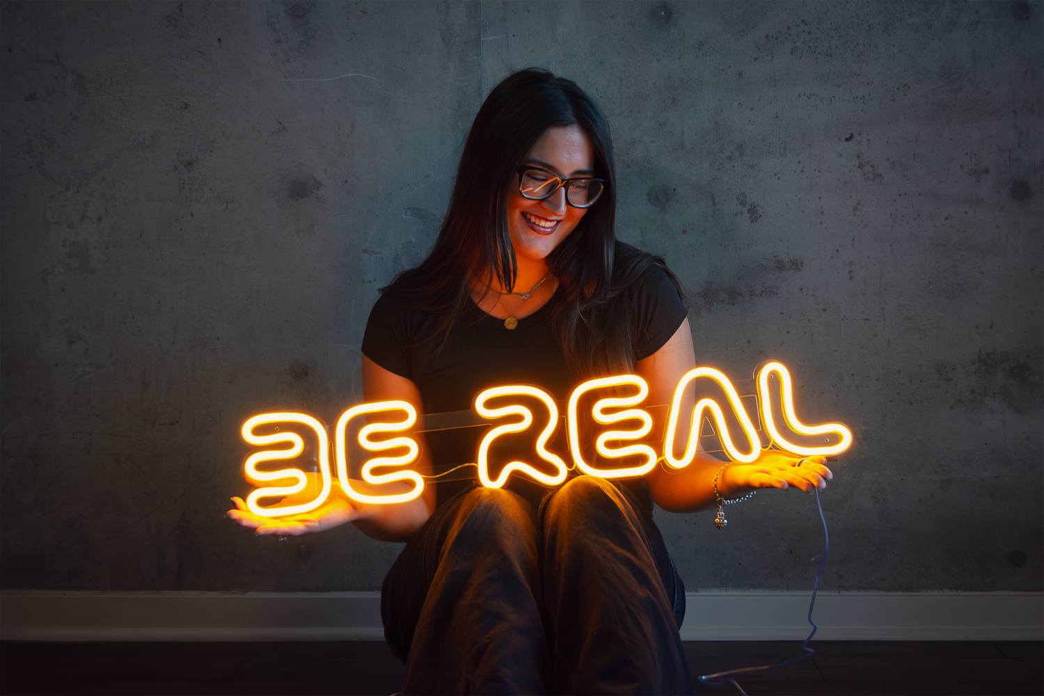 girl sitting while holding a yellow neon sign that says "be real"